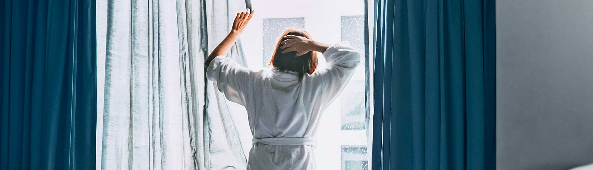ATLANTIC Hotels – Aufstehen und wohlfühlen Person in bathrobe opens curtains in a hotel room, daylight streams in, laptop on unmade bed.