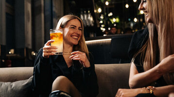 Zwei lachende Frauen in einer stilvollen Hotelbar, eine hält ein Getränk.