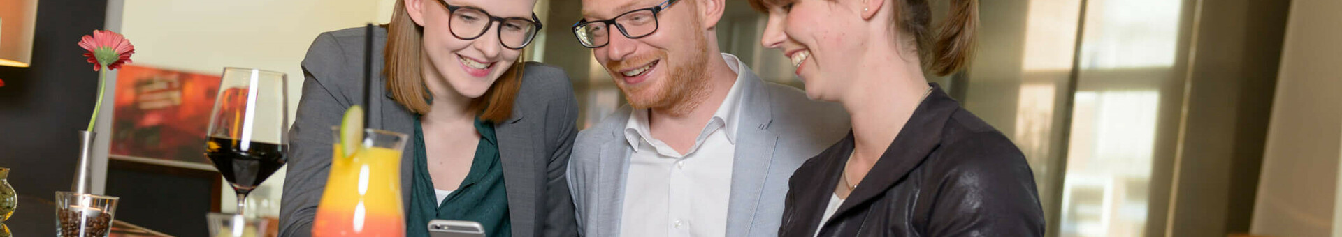 Three people laughing at a hotel bar, looking at a smartphone; drinks and candle on the counter.