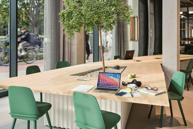 Modern work area in the ATLANTIC Hotel Münster with large wooden table, green chairs and laptop, surrounded by a glass front.