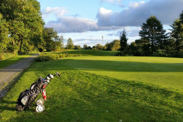 Golfplatz mit Golftasche am Rand, umgeben von grünen Bäumen und Rasen unter blauem Himmel mit Wolken.