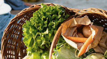 Paar sitzt auf Holzsteg am Wasser, neben ihnen ein Korb mit frischem Gemüse.