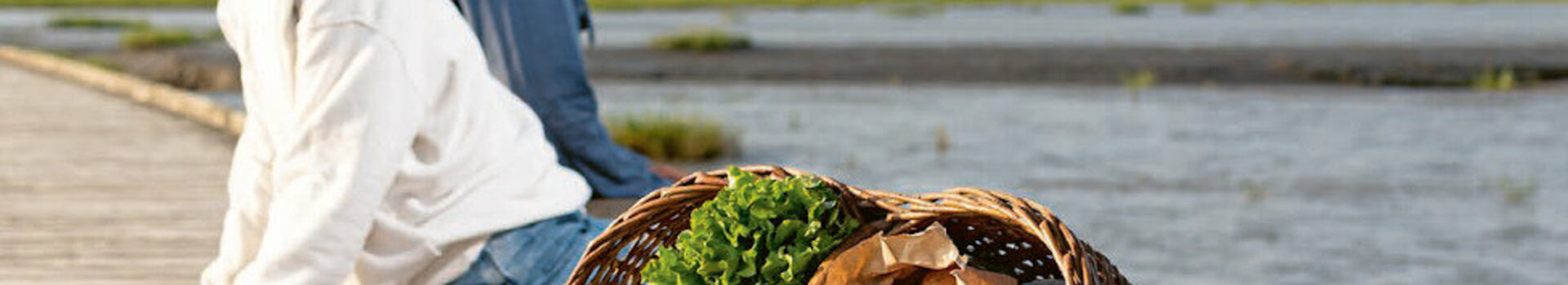 Watt'n Cookbook at the ATLANTIC Hotel Wilhelmshaven Couple sitting on a wooden jetty by the water, a basket of fresh vegetables next to them.
