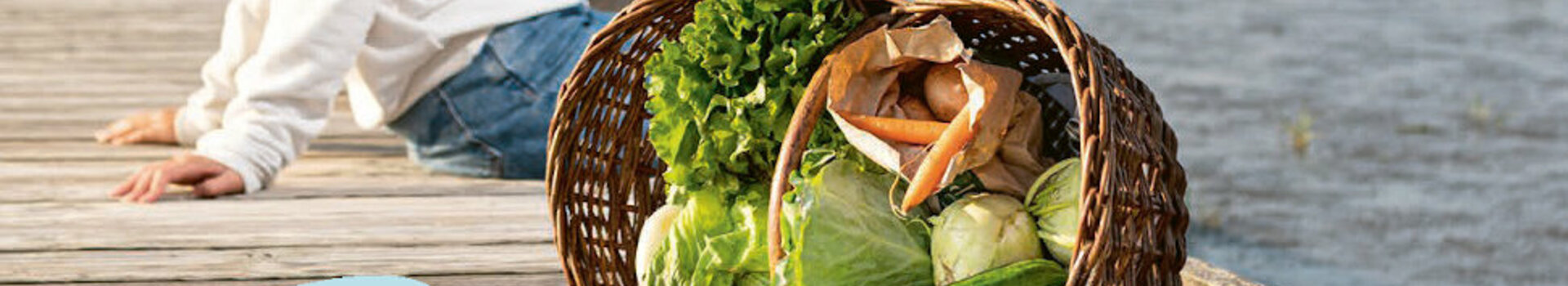 Watt´n Kochbuch im ATLANTIC Hotel Wilhelmshaven Paar sitzt auf Holzsteg am Wasser, neben ihnen ein Korb mit frischem Gemüse.