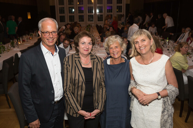 Four smiling people in evening dress at a festive dinner with set tables in the background.