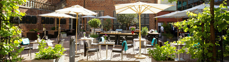 Der Hofgarten mit grünen Bäumen, Steinterrasse, Sonnenschirmen und Sitzgruppen im Sonnenlicht.