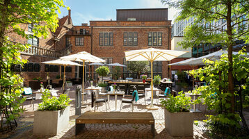 The courtyard garden with its green trees, stone terrace, parasols and seating areas bathed in sunlight.