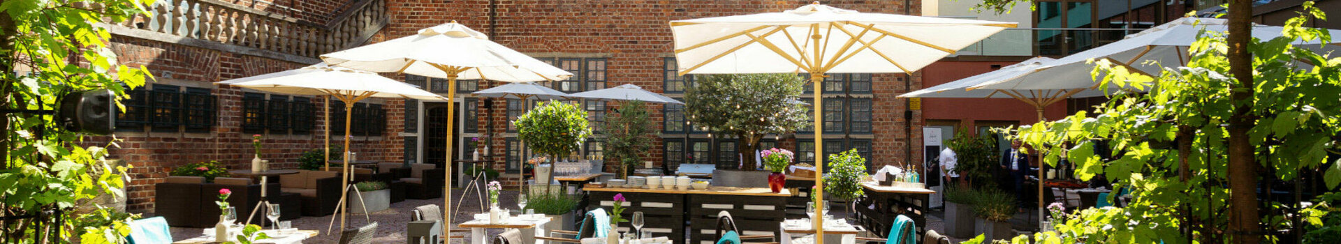 The courtyard garden with its green trees, stone terrace, parasols and seating areas bathed in sunlight.