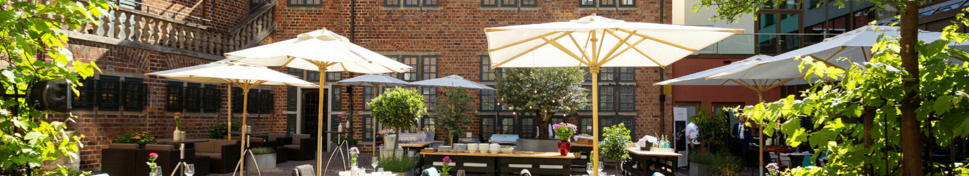 Der Hofgarten mit grünen Bäumen, Steinterrasse, Sonnenschirmen und Sitzgruppen im Sonnenlicht.