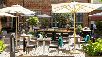 Der Hofgarten mit grünen Bäumen, Steinterrasse, Sonnenschirmen und Sitzgruppen im Sonnenlicht.