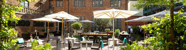 The courtyard garden with its green trees, stone terrace, parasols and seating areas bathed in sunlight.