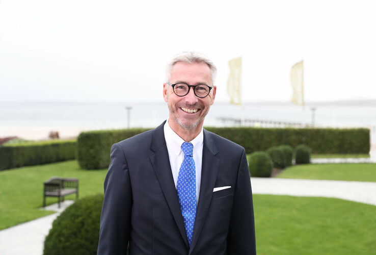 Ein lächelnder Mann im Anzug steht im Garten des ATLANTIC Grand Hotels Travemünde mit Meerblick im Hintergrund.
