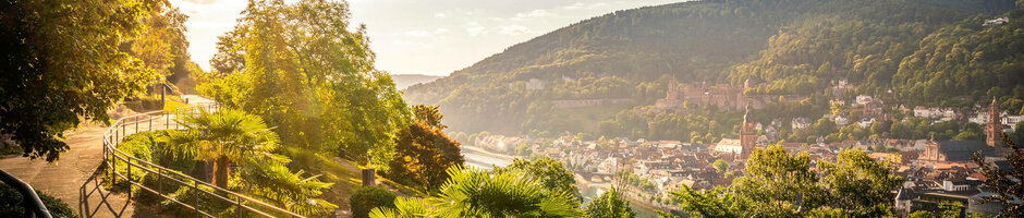 ATLANTIC Hotel Heidelberg Rahmenprogramm Philosophenweg Sonniger Panoramaweg mit Blick auf eine Stadt und bewaldete Hügel im Hintergrund.