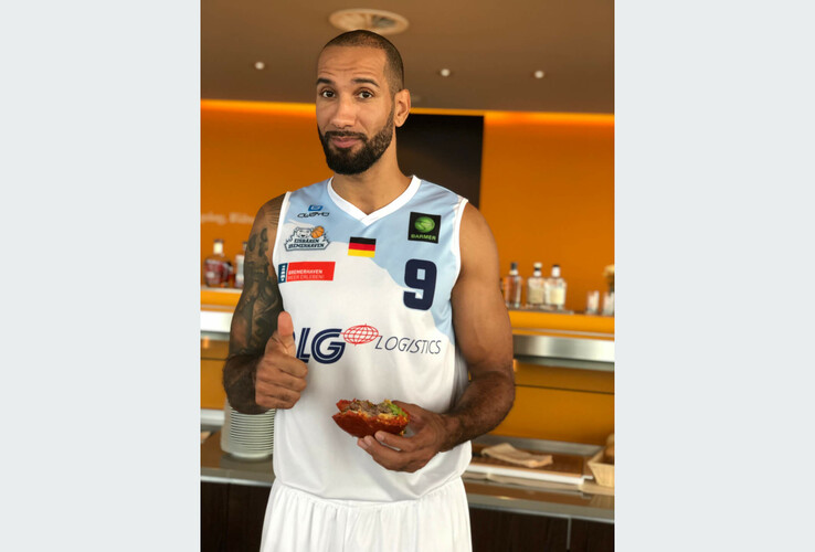 Basketball player in jersey eating a burger and giving a thumbs up in a hotel restaurant.