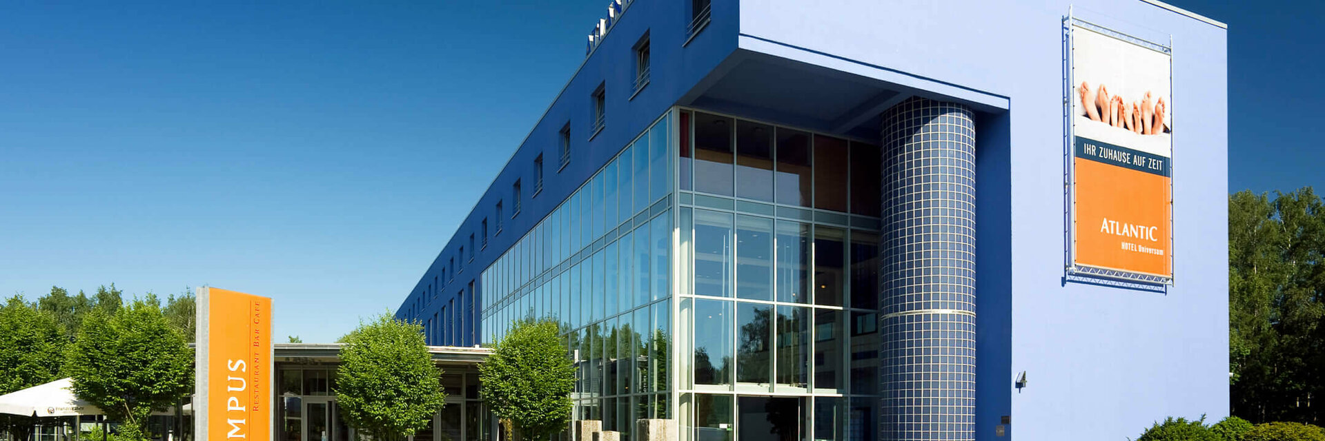ATLANTIC Hotel Universum in Bremen: Modern, blue building with glass façade, surrounded by manicured lawns and trees.