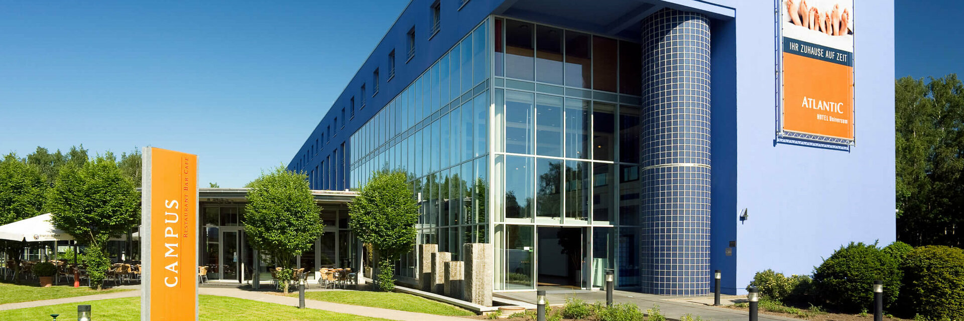 ATLANTIC Hotel Universum in Bremen: Modernes, blaues Gebäude mit Glasfassade, umgeben von gepflegtem Rasen und Bäumen.