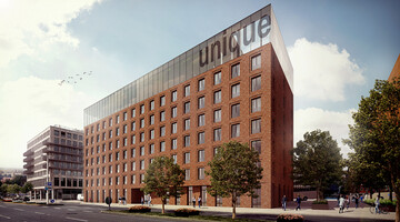 Modern hotel building with red brick façade, large windows and glass roof, surrounded by trees and pedestrians.