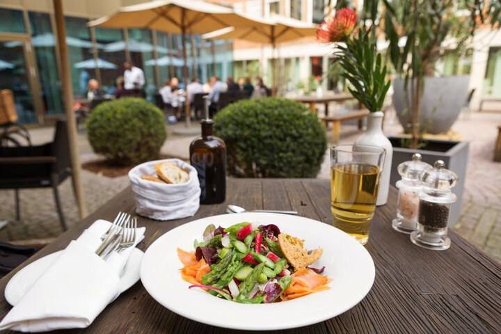Sommerlicher Salat mit Spargel und Lachs im Hofgarten des ATLANTIC Grand Hotel Bremen  Salat mit Lachs auf Holztisch im ATLANTIC Grand Hotel Bremen, Terrasse mit Gästen im Hintergrund.