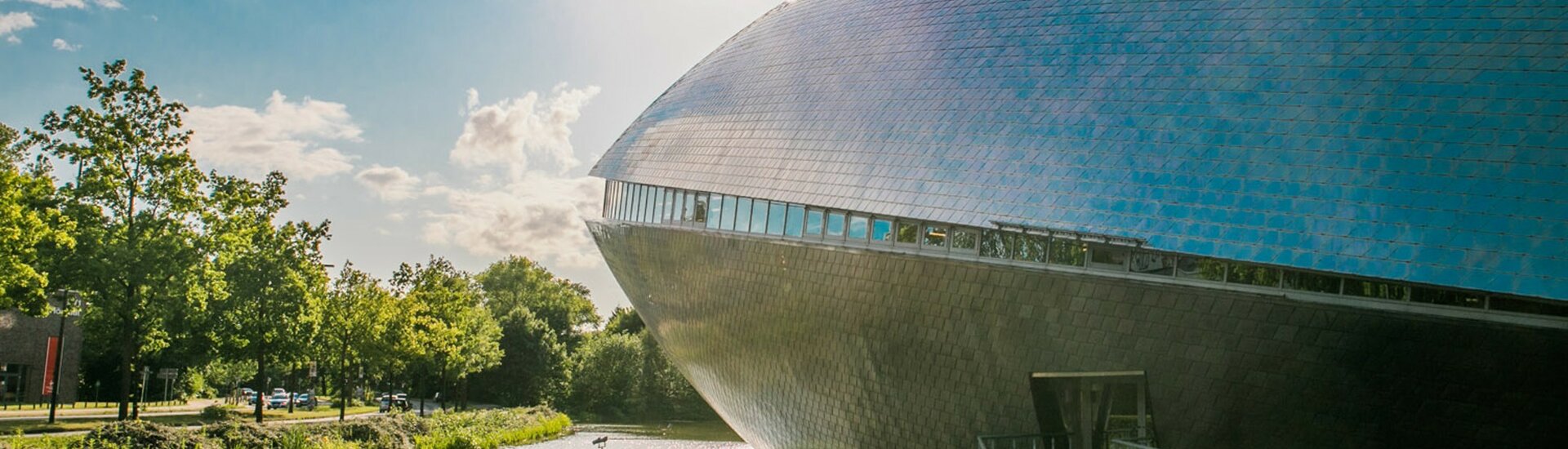 Außenansicht des Universum Bremen Moderne Architektur bei Sonnenschein: Rundes, metallisches Gebäude neben einem Wassergraben, umgeben von Bäumen.
