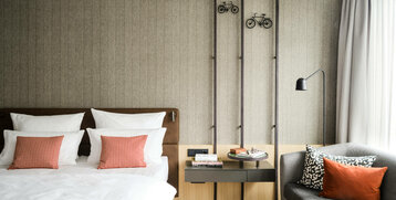 Modernes Hotelzimmer mit Doppelbett, grauem Sofa, roten Kissen und stilvoller Wanddekoration im ATLANTIC Hotel Münster.