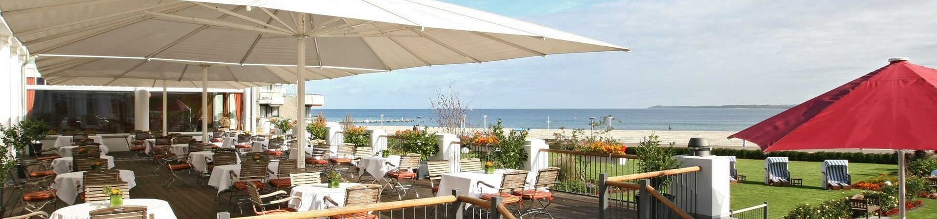 Hotelterrasse mit Tischen und Stühlen, Blick auf die Ostsee, Sonnenschirme, gepflegter Garten mit Strandkörben.