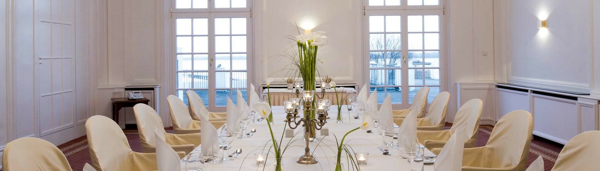 Eleganter, weißer Speisesaal mit gedecktem Tisch, Kerzen und Blumen, Kristallleuchter an der Decke, Blick aufs Wasser.