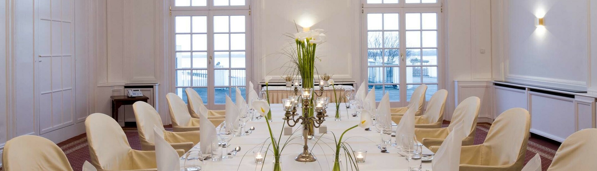 Elegant, white dining room with table setting, candles and flowers, crystal chandelier on the ceiling, view of the water.