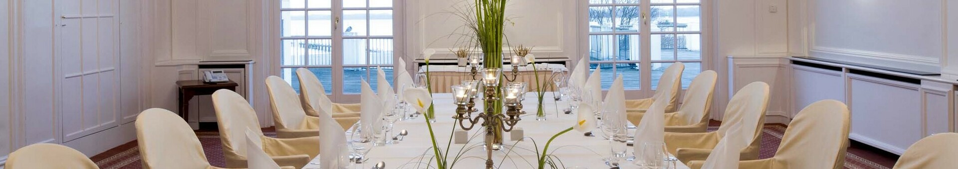 Veranstaltungsraum Kiel Eleganter, weißer Speisesaal mit gedecktem Tisch, Kerzen und Blumen, Kristallleuchter an der Decke, Blick aufs Wasser.