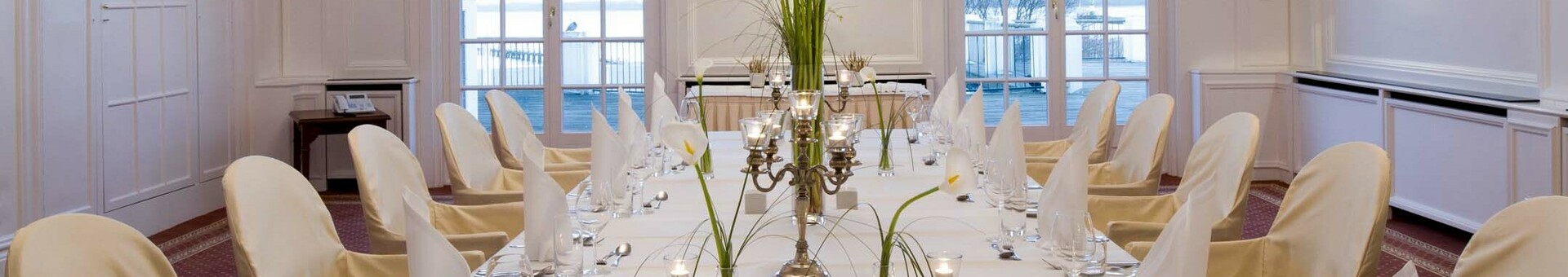 Elegant, white dining room with table setting, candles and flowers, crystal chandelier on the ceiling, view of the water.