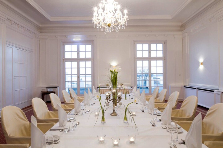 Veranstaltungsraum Kiel Eleganter, weißer Speisesaal mit gedecktem Tisch, Kerzen und Blumen, Kristallleuchter an der Decke, Blick aufs Wasser.