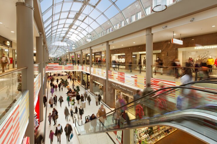 Modern shopping center with glass roof, escalators and stores on two floors, full of people.