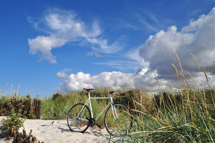 Fahrrad auf sandigem Pfad zwischen Dünen und Gras, unter blauem Himmel mit weißen Wolken.