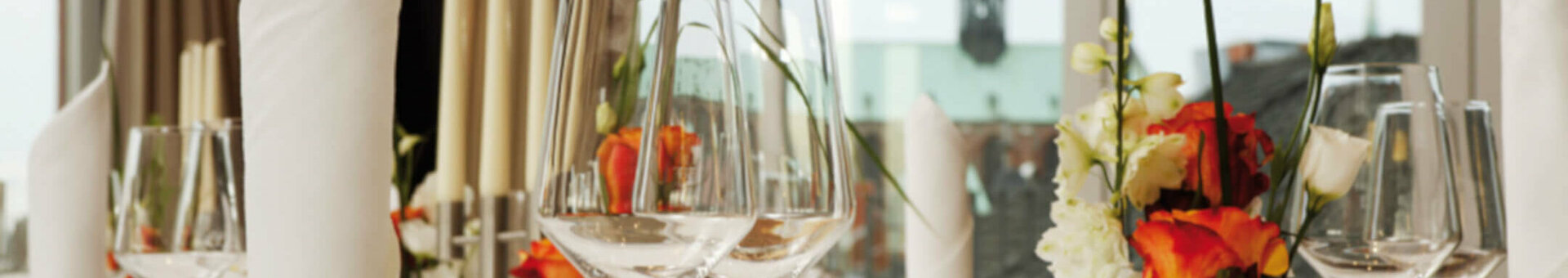 Elegant table setting at the ATLANTIC Hotel Lübeck with flowers, glasses and cutlery in front of large windows.
