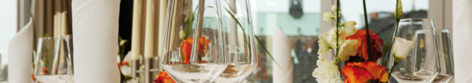 Eleganter, gedeckter Tisch im ATLANTIC Hotel Lübeck mit Blumen, Gläsern und Besteck vor großen Fenstern.