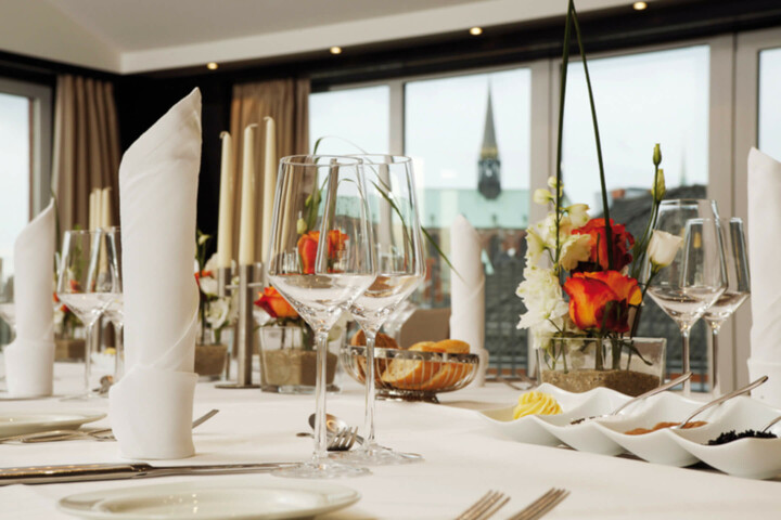 Eleganter, gedeckter Tisch im ATLANTIC Hotel Lübeck mit Blumen, Gläsern und Besteck vor großen Fenstern.