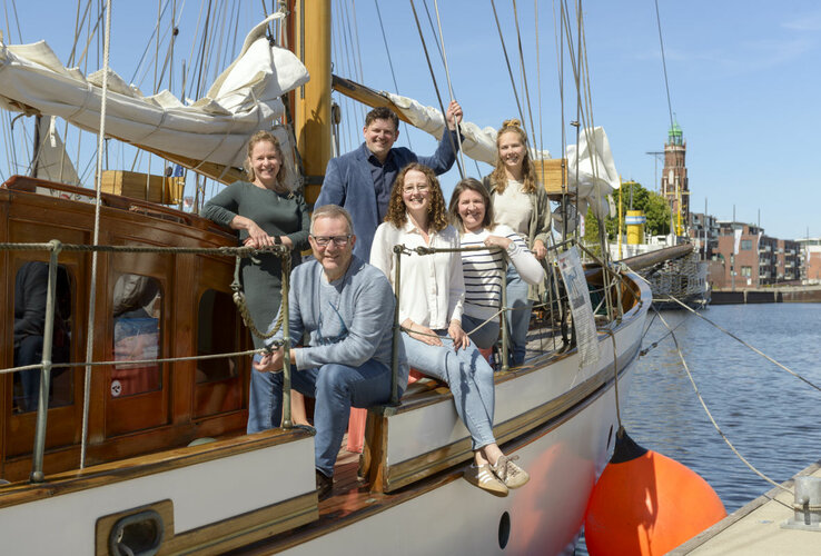6 Personen sitzen und stehen auf einem kleinen Segelschiff und lächeln bei sonnigem Wetter in Richtung der Kamera
