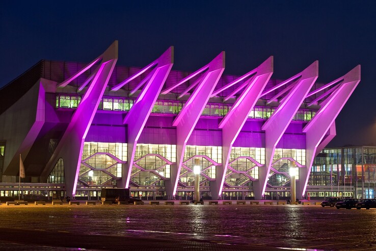 Modernes Gebäude mit markanten, pink beleuchteten Streben und großen Glasfenstern bei Nacht.