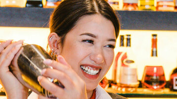 Foto einer Mitarbeiterin hinter der Bar. Sie schüttelt den Cocktailshaker und lacht dabei. Im Hintergrund eine Wand voller Getränkeflaschen