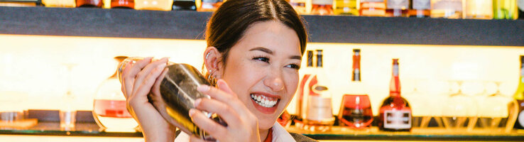 Foto einer Mitarbeiterin hinter der Bar. Sie schüttelt den Cocktailshaker und lacht dabei. Im Hintergrund eine Wand voller Getränkeflaschen