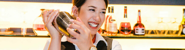 Photo of a female employee behind the bar. She is shaking the cocktail shaker and laughing. In the background, a wall full of drinks bottles.