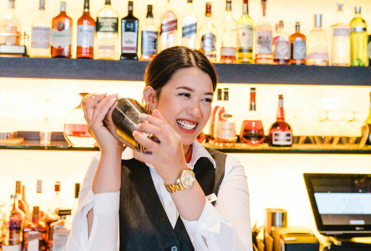 Photo of a female employee behind the bar. She is shaking the cocktail shaker and laughing. In the background, a wall full of drinks bottles.