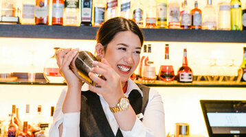 Photo of a female employee behind the bar. She is shaking the cocktail shaker and laughing. In the background, a wall full of drinks bottles.