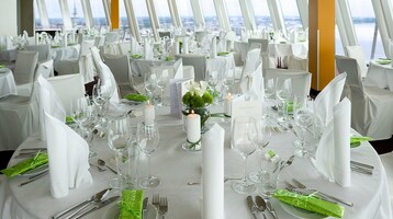 Eleganter Bankettsaal mit runden, weiß gedeckten Tischen, grünen Akzenten und Blick auf die Stadt durch große Fenster.