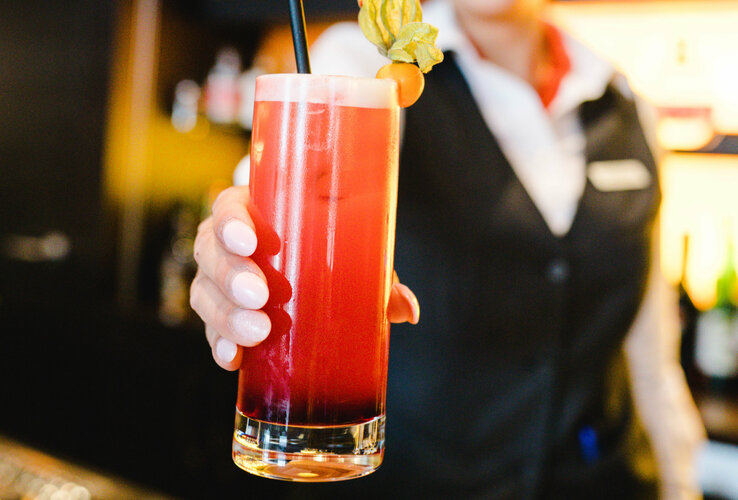 Close-up of a red cocktail with decoration, blurred in the background is the employee holding it up to the camera.