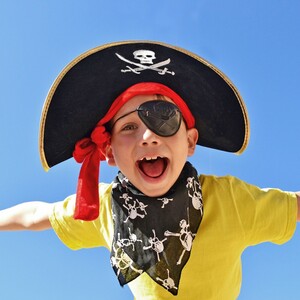 Kind in Piratenkostüm mit Augenklappe und Hut lacht fröhlich vor blauem Himmel.