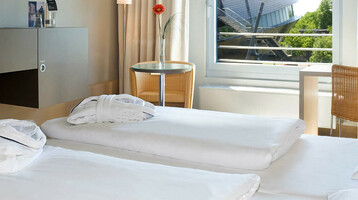 Helles Hotelzimmer mit zwei Betten, Schreibtisch, Fernseher und großem Fenster mit Blick auf grüne Bäume.