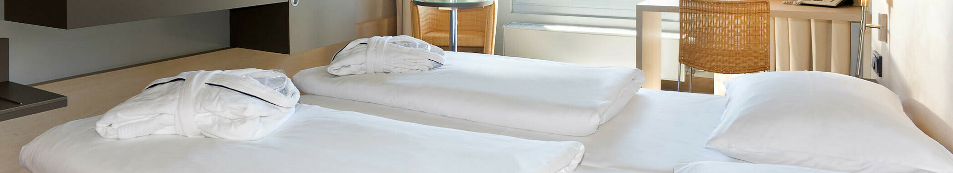 Business Zimmer im ATLANTIC Hotel Universum Helles Hotelzimmer mit zwei Betten, Schreibtisch, Fernseher und großem Fenster mit Blick auf grüne Bäume.