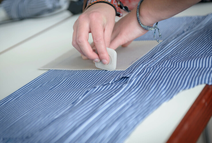 Person markiert Stoff mit Kreide für Schneiderei, Detailaufnahme von Händen und blau-weiß gestreiftem Stoff.