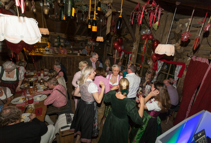People in traditional costume celebrate in a rustic, cozy atmosphere with decorations and drinks.