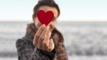 Person hält ein rotes Herz vor unscharfem Meereshintergrund.